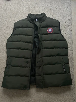 Mörkgrön dunväst från Canada Goose - Säljer en mörkgrön dunväst från Canada Goose med klassisk logotyp på bröstet. Västen har quiltad design, hög krage och praktiska fickor med knappar och dragkedja. Perfekt för kyliga dagar och enkel att matcha med din garderob.