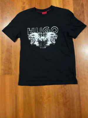 Svart HUGO t-shirt med tryck - Svart t-shirt från HUGO med ett stort, grafiskt tryck av två ritade varghuvuden och HUGO-loggan i vitt på bröstet. Klassisk rund hals och korta ärmar. Materialet är mjuk bomull som känns skönt mot huden. Perfekt för dig som gillar streetwear och vill sticka ut.