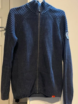 Marinblå stickad kofta från Sail Racing - Säljer en marinblå stickad kofta från Sail Racing med grov ribbstickning och hel dragkedja framtill. Koftan har hög krage, lång ärm och ett snyggt expeditionsmärke på ena ärmen. Perfekt för kyliga dagar och riktigt skön att ha på sig.