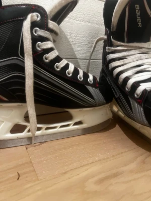 Svart och vit Bauer skridskoväst - Säljer en svart och vit väst från Bauer med coola vita snören och detaljer i grått och rött. Västen har en sportig look med högt skaft och tydliga Bauer-loggor. Materialet är syntet och designen är gjord i Kanada. Perfekt för dig som gillar att sticka ut på isen.