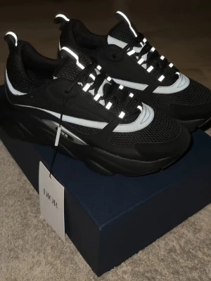 Svarta Dior b22 - Säljer ett par svarta Dior sneakers med feta reflexdetaljer och vita inslag. Skorna har chunky sula, meshpartier och snygga snörningar. Märket syns på sidan och plösen. Perfekt för dig som vill sticka ut med exklusiva och trendiga sneakers.