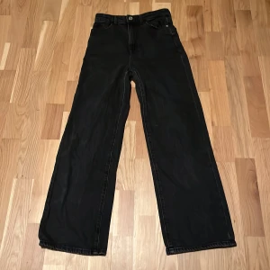 Svarta wide jeans från Lindex - Svarta jeans från Lindex med breda ben och klassisk femficksdesign. Byxorna har hög midja, bälteshällor och knappstängning fram. Materialet är jeans och färgen är helsvart, vilket ger en clean och stilren look. Använda flertal gånger men dem har hållit sitt skick.