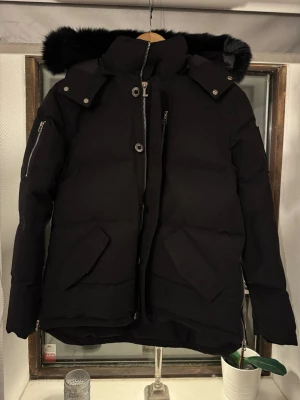 Svart dunjacka från Moose Knuckles - Säljer en svart dunjacka från Moose Knuckles med stor huva och fluffig pälsdetalj. Jackan har flera fickor med lock och dragkedja, samt snygga metalldetaljer. Jackan är ny, endast provad!