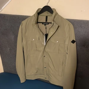 Beige vindjacka från J.Lindeberg - Stilren olivgrön vindjacka från J.Lindeberg med silverfärgade knappar och dragkedja. Jackan har fyra framfickor med lock, hög krage och ett svart märke på ärmen. Materialet är slätt och syntetiskt, perfekt för vår och höst. Passformen är normal och jackan är långärmad och den har en inbyggd luva! Jackan är köpt för 3200 den har aldrig använts så den är för stor på mig!  Priset kommer inte diskuteras! 