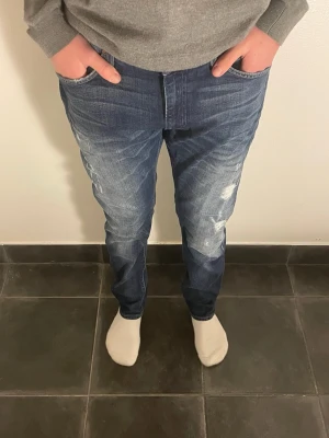 Mörkblå Lee jeans med slitningar 180-187 - Säljer ett par mörkblå jeans från Lee med snygga slitningar och tvättade detaljer på benen. Modellen har raka ben, klassiska fem ficks modell.