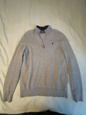 Grå half zip-tröja från Morris - Snygg grå tröja från Gant med half zip och liten broderad logga på bröstet. Tröjan har lång ärm och ribbade muddar. Perfekt för lager-på-lager och funkar året runt. Materialet känns mjukt och är troligtvis bomull eller ullblandning. Litet hål vid armhålan men inte något som inte går att fixa.