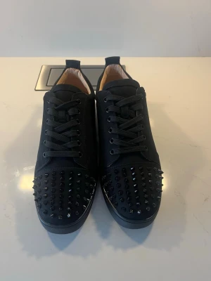 Christian Louboutin svarta sneakers med nitar - Säljer ett par svarta sneakers från Christian Louboutin med coola svarta nitar på tån och klassisk röd sula. Skorna har snörning och är gjorda i skinn med en rund tå. Medföljer dustbag och kvitto. Perfekt för dig som vill sticka ut med lyxiga detaljer.