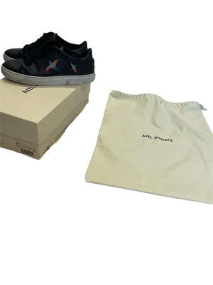 Svarta sneakers Axel Arigato  - Svarta sneakers från Axel Arigato med färgglada fågelmotiv på sidorna. Skorna har snörning, rund tå och vit platt sula. Tillverkade i skinn och kommer med originalkartong och dustbag. Perfekt för dig som gillar stilrena och unika detaljer.