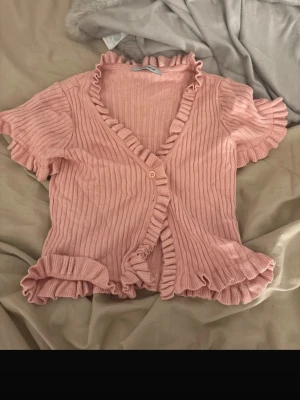 Rosa ribbad blus med volang - Supersöt ljusrosa ribbad blus från Stradivarius med volangdetaljer längs kanterna och ärmsluten. Kortärmad och croppad modell med v-ringning och knappar framtill. Perfekt för vår och sommar när du vill ha en gullig och trendig look.