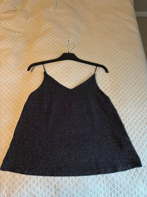 Svart glittrig linnetopp med smala axelband - Säljer en svart glittrig linnetopp med smala axelband och v-ringad hals. Toppen har en lätt A-linjeformad siluett och är tillverkad i ett syntetmaterial som skimrar snyggt i ljuset. Perfekt för att lyfta din outfit med lite extra glitter.