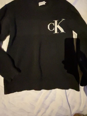 Svart stickad tröja Calvin Klein - Svart stickad tröja från Calvin Klein Jeans med stort vitt CK-tryck på bröstet.  S/M 