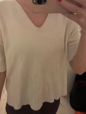 Tröja - Mysig beige stickad tröja med v-ringning och korta ärmar. Tröjan har ribbade detaljer vid ärmslut och nederkant, vilket ger en snygg och enkel look. Perfekt att slänga på sig när det är lite kyligt ute.