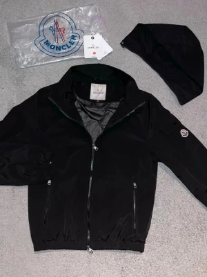 Svart Moncler vindjacka med huva - Svart vindjacka från Moncler med avtagbar huva och två sidofickor med dragkedja. Jackan har elastiska muddar vid ärmslut och nederkant samt Moncler-logga på ärmen. Perfekt för dig som gillar stilren och funktionell design.