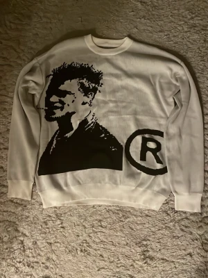 Vit Joy Divizn sweatshirt - Säljer en vit joy divizn sweatshirt med ett stort svart grafiskt tryck av Brad Pitts ansikte och ett R-symbol bredvid. Tröjan är flared och har rund hals, tillverkad i mjukt material och passar perfekt för dig som gillar streetwear och unika prints.