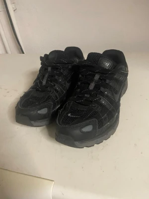 Svarta Nike P-6000 - Säljer ett par helsvarta Nike sneakers med cool mesh och detaljer i syntet och skinn. Skorna har snörning, rund tå och en chunky sula som ger grym streetkänsla. Perfekta för dig som gillar sportig stil och vill ha något som funkar till det mesta.