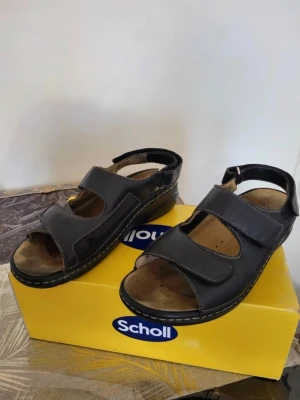 Svarta Scholl sandaler med kardborre 38 - Scholl sandaler i svart skinn med rund tå och platt sula. De har två breda kardborreband över foten och ett runt hälen för enkel justering. Sulan är ergonomiskt utformad och innersulan är beige med små perforeringar för ventilation. Perfekt för varma dagar.