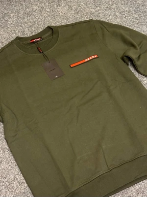 Olivgrön sweatshirt från Prada - Snygg olivgrön sweatshirt från Prada med rund hals och lång ärm. Tröjan har en röd detalj med Prada-logga på bröstet och ribbade muddar vid ärmslut och nederkant. Perfekt för dig som gillar stilrena designer och exklusiva märken.