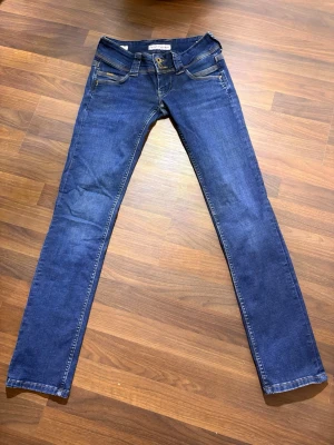 Pepe Jeans låg midja  - Snygga blå lågmidjade jeans från Pepe Jeans med slim straight passform och klassisk femficksdesign. Jeansen har mörkblå tvätt, kontrastsömmar och läderpatch bak i midjan. Materialet är jeans med Wiser Wash-teknik för en fräsch look. Storlek 24/32 skulle nog säga att det motsvarar stl S 