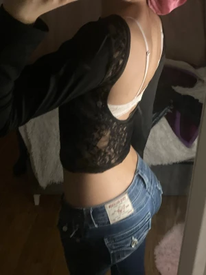 Svart croppad långärmad spetsblus - Säljer en svart croppad blus med lång ärm och öppen rygg. Toppen har genomskinliga spetsdetaljer på ryggen och sidorna, vilket ger en snygg och edgy look. Perfekt att styla med jeans eller kjol för en trendig outfit.