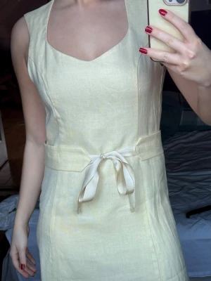 Ljus ärmlös klänning med knyt - Supersnygg ljus klänning i linne med bred midjedetalj och knytband framtill. Modellen är ärmlös och har en mjukt skuren halsringning. Perfekt för dig som gillar minimalistisk stil och vill ha något som känns fräscht och somrigt. Ger lite 80-tals vibe vilket jag älskar. 