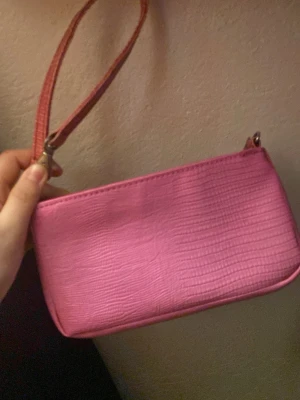 Rosa axelväska från Estée Lauder - Säljer en snygg rosa axelväska från Estée Lauder med reptilmönstrad yta och dragkedja. Väskan har en kompakt och rektangulär form med matchande axelrem. Perfekt för dig som gillar färg och vill ha något som sticker ut.