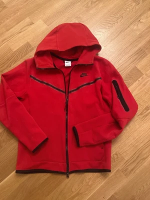 Nike Teck Fleece Röd - strlk S - bra skick - Nike Tech Fleece-kofta i röd färg, storlek S. Bra skick. Det finns ett pytte litet hål i fickan, knappt synligt och inget som påverkar användningen. I övrigt inga fläckar eller större slitage. Mjuk, varm och bekväm – perfekt till vardag och streetwear.
