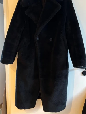 Svart teddy/fuskpäls-kappa från H&M – oversized - Lång svart kappa i mjuk teddy/fuskpäls från H&M i storlek XS. Klassisk och tidlös modell med bred krage och dubbelknäppning. Varm och skön, enkel att styla både till vardag och mer uppklädda tillfällen. Modellen är stor i storleken och har en tydlig oversized passform.  Fint skick. Väl omhändertagen. XS men stor i storleken, passar även S/Mž beroende på önskad passform.