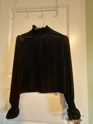 Svart sammetsblus med volangkrage - Säljer en svart blus i mjuk sammet med hög volangkrage och långa puffärmar med volangdetalj vid ärmslutet från Mango. Blusen har en elegant och lite vintage vibe, perfekt för dig som gillar unika detaljer och skön känsla. Använt endast 2-3 ggr.