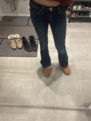True Religion blå bootcut jeans - Säljer ett par mörkblå jeans från True Religion med bootcut passform och snygga detaljer på bakfickorna. Jeansen har låg midja, klassiska kontrastsömmar och stora knappar på fickorna. Materialet är jeans med en skön stretch. Perfekt för dig som gillar en cool och avslappnad stil.