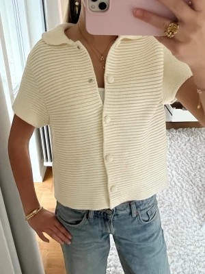 Vit stickad kofta från ZARA - Säljer en vit stickad kofta från ZARA i storlek M. Koftan har korta ärmar, krage och knappar framtill. Den är ribbad.