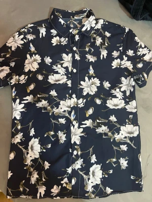  marinblå skjorta - Snygg kortärmad skjorta i marinblått med vita och beige blommor samt fjärilar. Skjortan har klassisk krage och knappar framtill. Perfekt för dig som gillar att sticka ut med ett fräscht blommigt mönster. Materialet känns lätt och skönt mot huden. Den är väldigt fin på en  sommarkväll du kan returnera direkt tillbaka om du inte gillar. Passar M och S. 