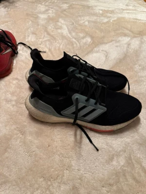 Adidas Ultraboost svarta sneakers - Svarta Adidas Ultraboost sneakers med grå detaljer och klassiska tre ränder på sidan. Skorna har snörning, stickad ovandel och en vit boost-sula med inslag av rött undertill. Väldigt bra skick