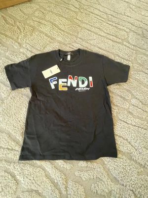 Svart Fendi t-shirt med färgglad logga - Cool svart t-shirt från Fendi med stor, färgglad logotyp på bröstet. T-shirten har rund hals och korta ärmar. Materialet är mjuk bomull och passformen är normal. Perfekt för dig som gillar streetstyle och vill sticka ut med designerplagg.