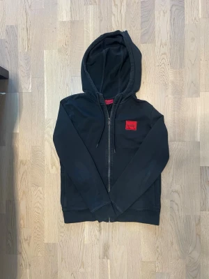 Hugo hoodie - Svart - Svart hoodie från Hugo med röd logotyp på bröstet. Tröjan har huva, hel dragkedja framtill och långa ärmar. Tillverkad i mjuk bomull, perfekt för en chill och stilren look. Den är i storlek Xs men skulle fungera perfekt för S också! Hör av dig vid intresse, pris kan diskuteras!