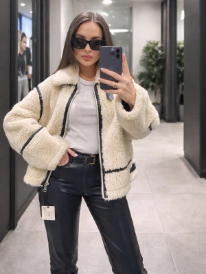 Teddy jacka från Zara  - Säljer en beige teddyjacka med svarta kontrastsömmar och dragkedja framtill. Jackan har en oversized passform, stor krage och är tillverkad i mjukt, fluffigt material som ger en cool och trendig look. Perfekt för att hålla sig varm och snygg under kyliga dagar.