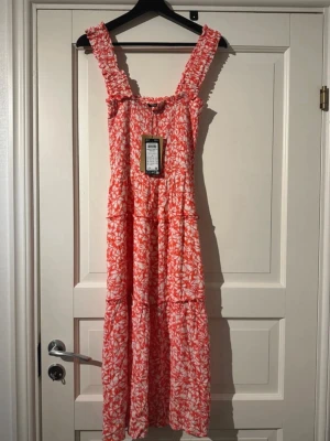 Blommig långklänning från Vero Moda - Säljer en superfin långklänning från Vero Moda i storlek XS. Klänningen har breda, rynkade axelband och ett rött och vitt blommigt mönster. Materialet är mjuk bomull och modellen är luftig med volanger. 