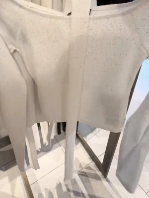 Glittrig vit långärmad topp med halsdetalj - Säljer en vit långärmad topp med glittriga detaljer och ett band runt halsen. Toppen har en tight passform och är tillverkad i ett mjukt, stretchigt material som känns bekvämt mot huden. Perfekt för dig som vill sticka ut lite extra.
