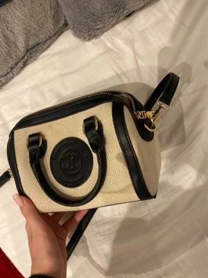 Beige och svart handväska - Snygg handväska  i beige canvas med svarta detaljer och handtag i skinn. Väskan har guldfärgade metalldetaljer och dragkedja upptill. Perfekt storlek för det viktigaste och har ett stilrent, klassiskt utseende med rund form.