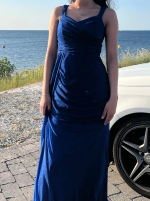 Marinblå glittrig långklänning - Säljer en marinblå långklänning med glittriga detaljer och breda axelband. Klänningen har draperad framsida och snygg v-ringning. Materialet är mjukt och faller fint längs kroppen, perfekt för speciella tillfällen.
