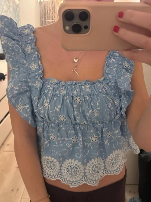 Blå broderad blus med volang - Supersöt blå blus med vita broderade blommor och volangdetaljer. Toppen har en fyrkantig ringning, rynkade axelband och en vågig, broderad nederkant. Perfekt för varma dagar och ger en romantisk vibe till outfiten.