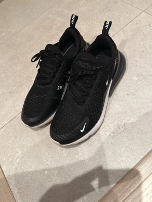 Nike Air Max 270 svarta sneakers - Svarta Nike Air Max 270 sneakers med vit sula och tydliga Air Max-detaljer. Skorna har snörning, mesh-överdel och en stor synlig luftkudde i hälen. Swoosh-logga i vitt på sidan och framtill. Perfekta för dig som gillar sportig och modern stil.