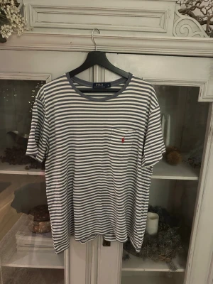 Randig t-shirt från Polo Ralph Lauren - Säljer en fräsch t-shirt från Polo Ralph Lauren i storlek XL. Den har blå och vita horisontella ränder, rund hals och en liten bröstficka med röd broderad logga. Materialet är mjuk bomull och passformen är normal. Perfekt för dig som gillar klassisk stil med en twist.