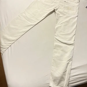 Vita raka jeans från J Lindeberg W32 L34 - Storlek W32 L34. Snygga vita jeans från Nudie med klassisk femficksdesign och raka ben. Byxorna har en enkel look med diskreta sömmar och en liten Nudie-etikett vid benslutet. Perfekt för dig som gillar stilrena och ljusa jeans. 