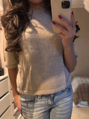 Beige stickad tröja med kort ärm - Mysig beige stickad tröja med korta ärmar och rund hals. Tröjan har en enkel och clean look som passar perfekt till jeans eller kjol. Materialet känns mjukt och fluffigt, vilket gör den extra skön att bära under kyligare dagar.