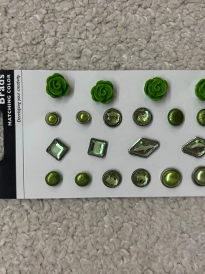 Gröna dekorativa sminkbrads - Säljer ett set med gröna dekorativa brads som kan användas för att pynta sminkförvaring eller sminkspeglar. Setet innehåller olika former som blommor, pärlor och glittriga stenar i grönt och silver. Perfekt för att ge din sminkhörna en personlig touch.