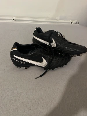 Nike Tiempo Light fotbollsskor svart - Säljer ett par svarta Nike Tiempo Light fotbollsskor i storlek 42 med vita och silvriga detaljer. Skorna har snörning, rund tå och platt sula med fasta dobbar. Tillverkade i skinn för bra känsla och komfort på planen. Snyggt Nike-märke på sidan och under sulan.