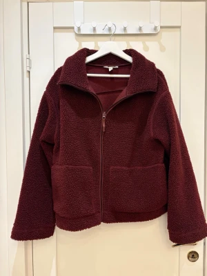 Vinröd teddyjacka med dragkedja - Mysig vinröd teddyjacka från H&M med två stora fickor framtill och hel dragkedja. Jackan har en bred krage och är tillverkad i fluffigt syntetmaterial som ger en cool och avslappnad vibe. Perfekt för kyliga dagar och enkel att styla till jeans eller leggings.