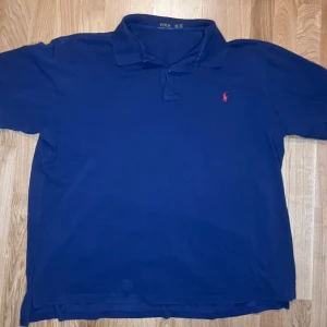 Blå pikétröja från Ralph Lauren - Klassisk marinblå pikétröja från Polo Ralph Lauren i storlek 3XL. Size taggen är dock overzised och stämmer inte riktigt. Den sitter mer som en XL Tröjan har korta ärmar, krage och knäppning framtill samt den ikoniska röda loggan broderad på bröstet. Tillverkad i mjuk bomull som känns skön mot huden.