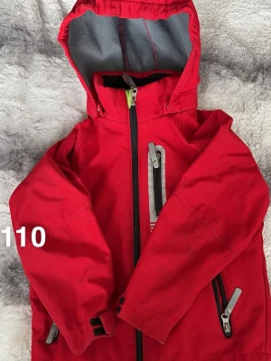 Röd jacka med huva, stl 110 - Säljer en röd jacka med huva och svart dragkedja. Jackan har flera fickor med dragkedjor och justerbara muddar vid ärmarna. 🌸 Fläckar. Storlek 110