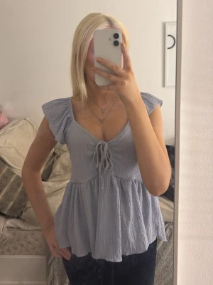 Ljusblå blus med knyt - Supersöt ljusblå blus från 157 med volangärmar och v-ringning. Toppen har knyt framtill och är tillverkad i ett tunt, krinklat bomullsliknande material som ger en luftig känsla. Perfekt till jeans eller kjol. Använd ett fåtal gånger.